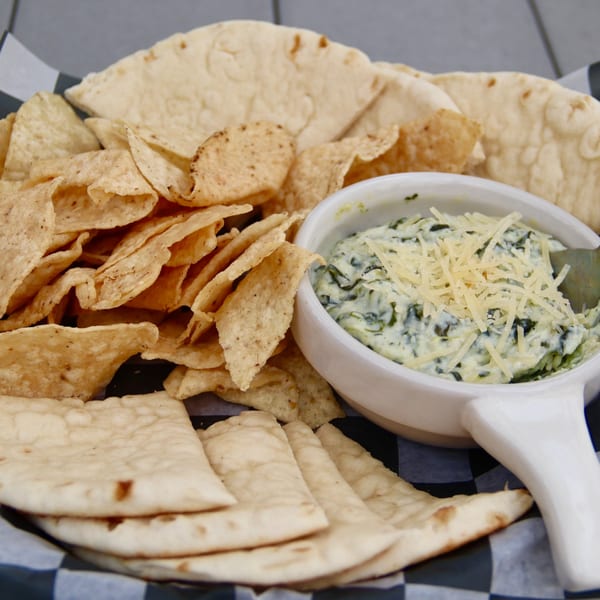 Spinach dip topped with shredded cheese, served with tortilla chips and pita bread in a basket.