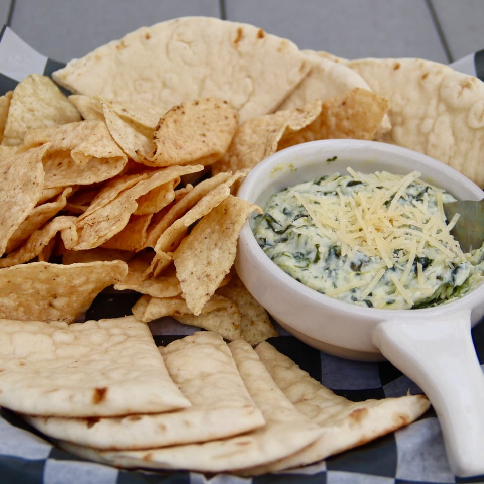 Spinach dip topped with shredded cheese, served with tortilla chips and pita bread in a basket.