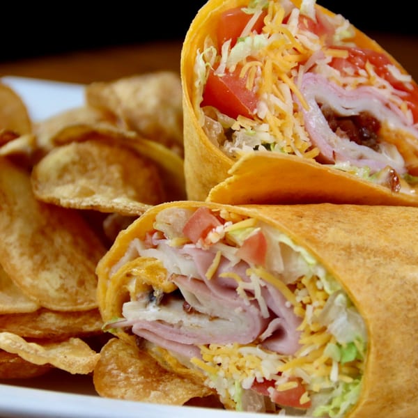 Two halves of a wrap filled with ham, cheese, lettuce, and tomato, served with potato chips.