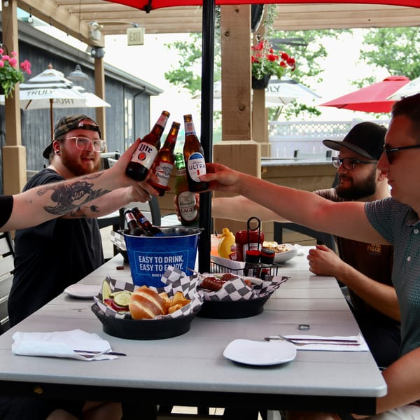 Four men toast beer bottles at an outdoor patio table with food and drinks.