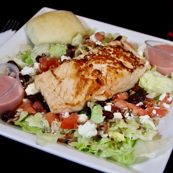 Grilled chicken salad with lettuce, tomatoes, feta, dried cranberries, sunflower seeds, and a bread roll.