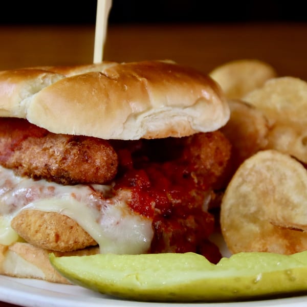 A Beckett's Burger Bar chicken parmesan sandwich with melted cheese, red sauce, and a pickle spear.