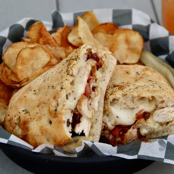 A chicken sandwich with melted cheese and sauce, served with potato chips and a pickle.